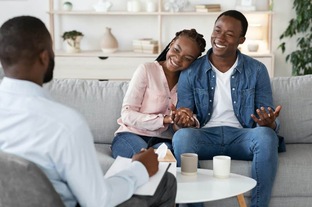 Family Counseling. Young Happy Black Couple Sitting On Couch At Psychotherapist's Office